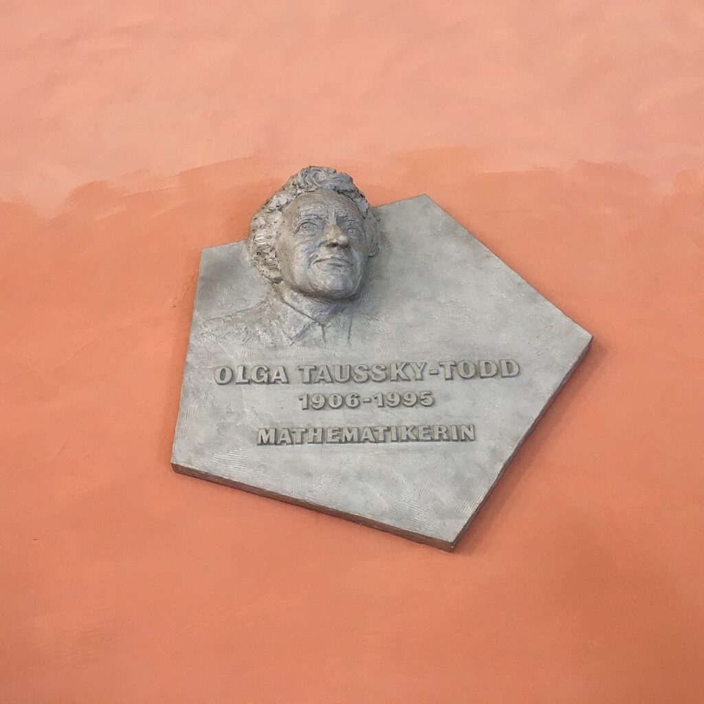 Wandrelief mit dem Porträt der Mathematikerin Olga Taussky-Todd, mit Namenszug und Lebensdaten, vor Orange-farbig gestrichener Wand.