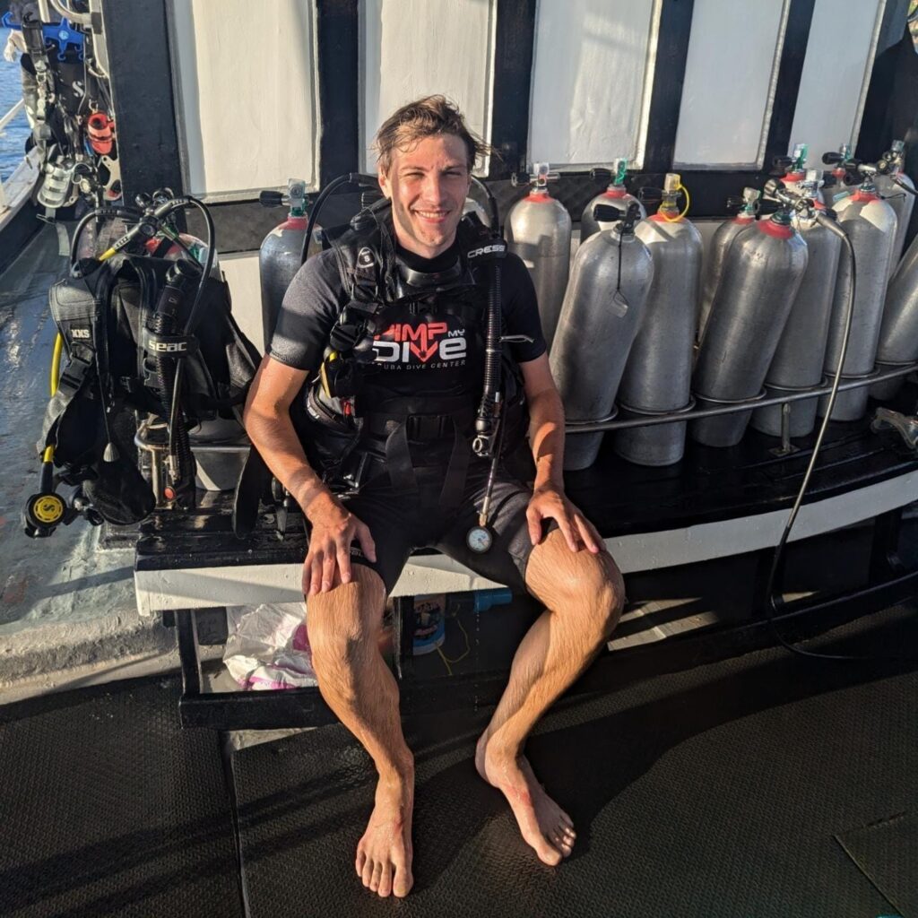 Auf dem Foto ist Florian sitzend mit einer Tauchausrüstung zu sehen. Neben ihm stehen Sauerstoffflaschen.