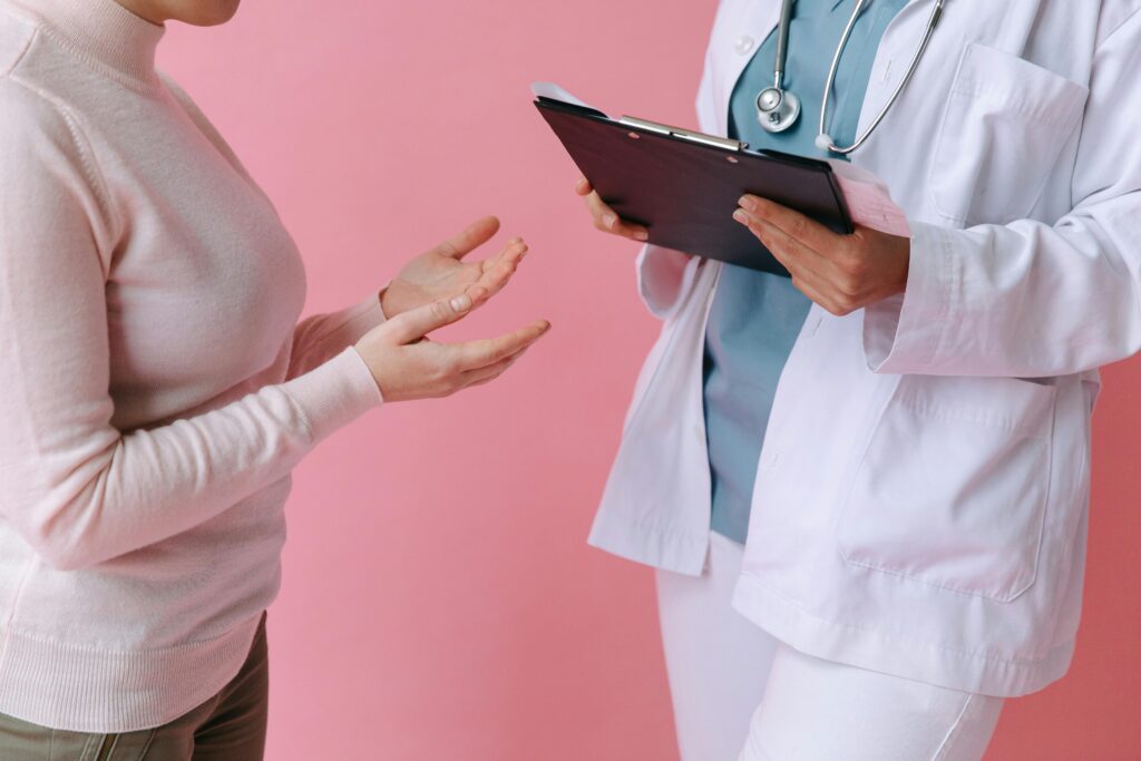 Patientin im rosa Rollkragenpullover spricht mit einer Ärztin in weißem Kittel und Stethoskop vor rosa Hintergrund – symbolisiert ein medizinisches Beratungsgespräch.