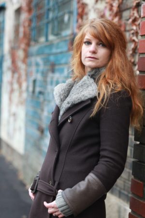 Porträt von Sandra Schäfer, die im Freien stehend an eine Wand gelehnt ist und in die Kamera blickt.