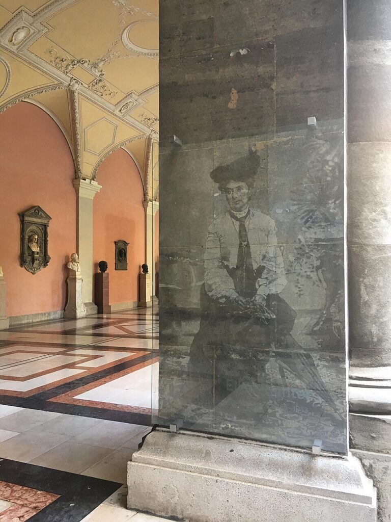 Transparentes Glasdenkmal mit einem historischen Porträt von Elise Richter im Arkadenhof der Universität Wien, vor einer Reihe männlicher Büsten.