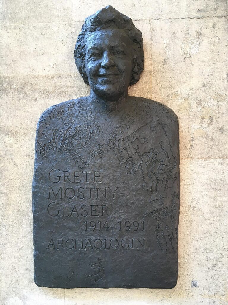 Reliefporträt der Archäologin Grete Mostny-Glaser mit Namenszug und Lebensdaten an einer Grauen Steinwand.