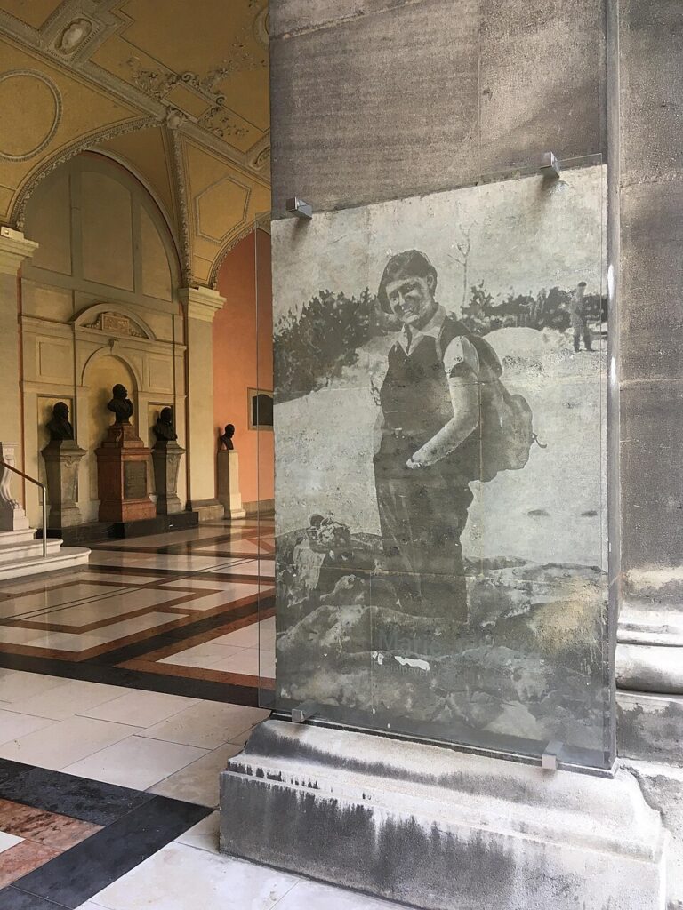 Glasinstallation mit einem historischen Porträt von Marie Jahoda im Arkadenhof der Universität Wien, umgeben von Säulen und Büsten.
