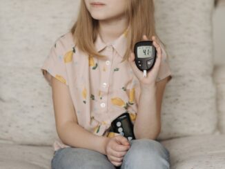 Ein Kind, das auf einer Couch sitzt und ein Blutzucker-Messgerät in die Kamera hält. Das gesamte Gesicht des Kindes ist nicht zu sehen, knapp oberhalb der Lippen ist das Bild abgeschnitten.