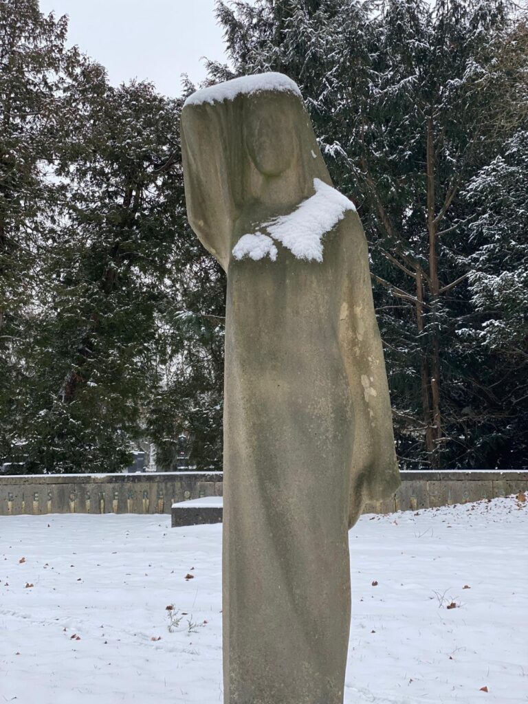 Steinerne Frauenfigur zugehörig zum Mahnmal gegen Faschismus am Wiener Zentralfriedhof, mit Hand am Kopf gestützt und langem Gewand, stehend in einer verschneiten Anlage.